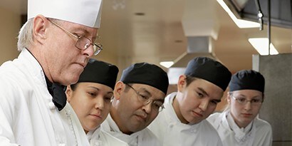 Um chef demonstra uma técnica de preparação de alimentos para quatro trabalhadores de cozinha que observam.
