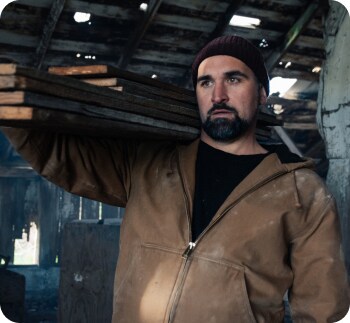 A bearded man carrying a stack of wood out of an old house.
