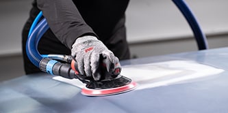 Trabalhador usando luvas de proteção operando uma lixadeira orbital 3M com um disco abrasivo para nivelar a superfície de um veículo.
