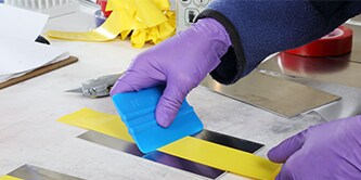 Gloved hands use a 3M™ Hand Applicator PA1 to smooth out a wide yellow masking tape applied to a metal rectangle
