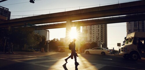 Distant view of a man crossing a crosswalk with the sun in the background.
