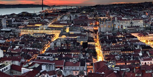 Vista panorâmica de Lisboa ao entardecer, com telhados avermelhados em primeiro plano, ruas iluminadas, edifícios históricos ao centro e o rio Tejo ao fundo com uma ponte suspensa sob céu alaranjado.