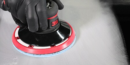 Uma pessoa usando luvas pretas usando uma lixadeira elétrica da marca 3M com um disco abrasivo azul para alisar uma superfície, criando um acabamento polido.
