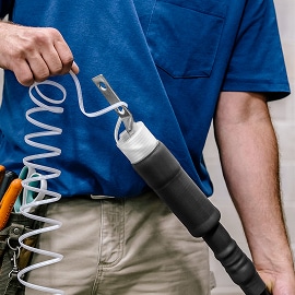 Trabalhador com camisa azul segurando uma mangueira de ar enrolada conectada a uma ferramenta pneumática preta, com cinto de ferramentas e calça cáqui visíveis.