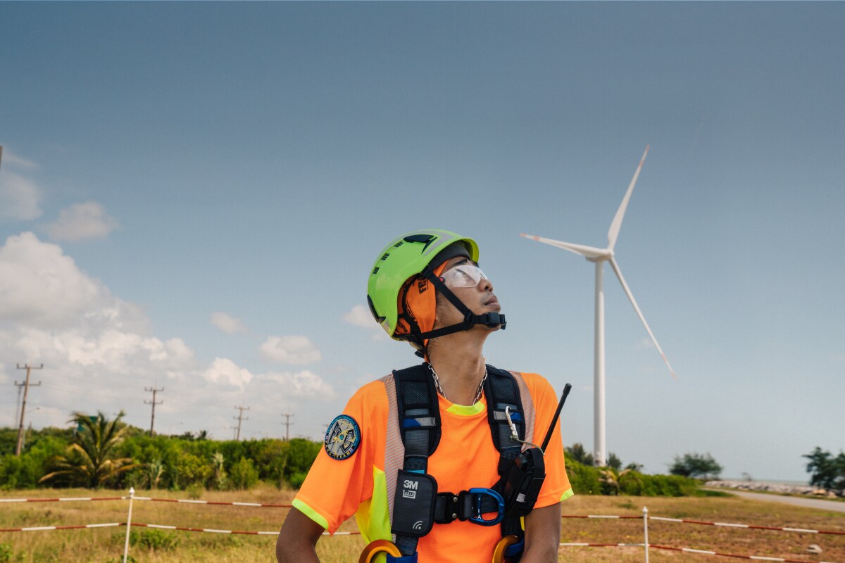 A man in protective gear looks up at a wind turbine.

