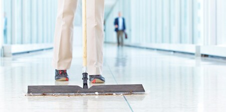 Imagem de um profissional de limpeza limpando com um rodo os resíduos do piso do corredor de uma escola.
