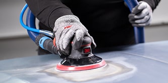 Trabalhador usando luvas de proteção utilizando uma lixadeira orbital da 3M com um disco abrasivo para nivelar a superfície de um veículo.
