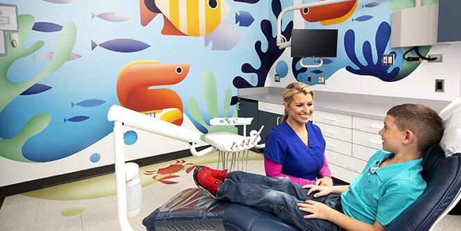 Criança na cadeira de dentista e técnica odontológica sorridente em um consultório de dentista, com uma ilustração vibrante de oceano impressa em Filmes Gráficos 3M.
