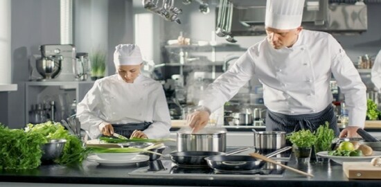 Três chefes preparam comida em uma cozinha comercial.
