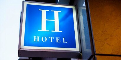 Sinal luminoso externo com uma grande letra H e “HOTEL” abaixo, fixado junto a um prédio.
