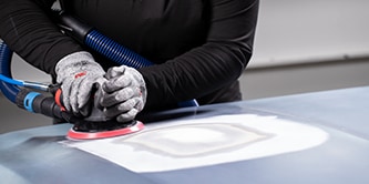 Trabalhador usando luvas de proteção utilizando uma lixadeira orbital com um disco abrasivo para nivelar uma área preparada da superfície de um veículo.
