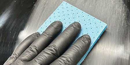 Uma pessoa vestindo uma luva preta segura uma boina abrasiva azul, lixando uma superfície escura para criar um acabamento liso e uniforme.
