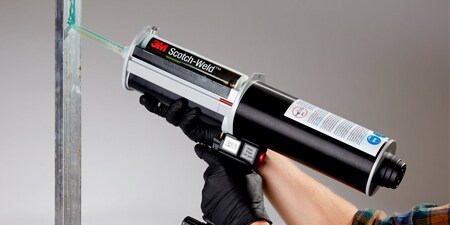 A gloved worker uses a pneumatic gun to apply a liquid adhesive to a vertical metal bar
