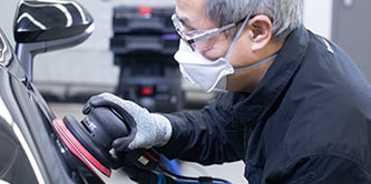 Trabalhador usando óculos de segurança, luvas e máscara de proteção enquanto utiliza uma politriz manual para preparar a superfície de um veículo.
