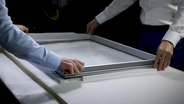 Fotograma de vídeo de dois trabalhadores colocando uma moldura em uma placa de vidro na qual foi aplicada a 3M™ Fita Extrudável VHB™ GP.
