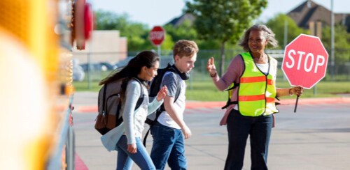 Young girl and boy crossing a crosswalk with the help of a school patrol officer holding a stop sign.
