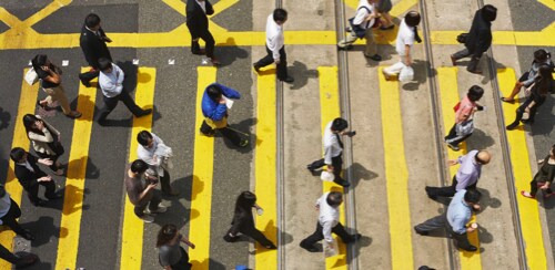 Large group of dispersed people crossing a yellow stripe painted crosswalk.
