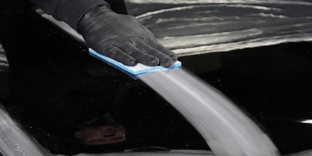 Uma pessoa vestindo luva preta usa uma boina abrasiva azul para lixar uma superfície preta brilhante, criando uma marca suave e fosca.
