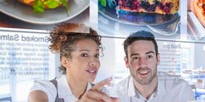Uma mulher e um homem sorridentes olham para um menu flutuante e colorido em um restaurante.
