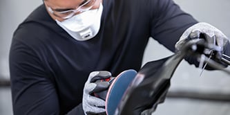 Trabalhador usando luvas de proteção operando uma lixadeira com um disco abrasivo azul sobre a superfície de um veículo.
