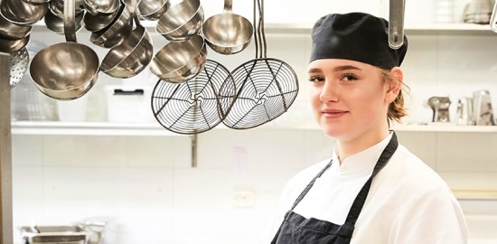 Uma trabalhadora da cozinha sorridente posando entre utensílios de cozinha pendurados.
