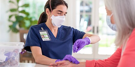 Clinician wearing a filtering facepiece respirator checking a patient’s pulse during a home visit
