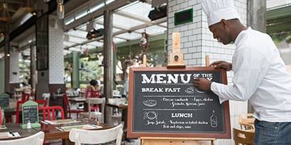 Chef atualizando o quadro de menu de giz exibido do lado de fora de um restaurante comercial.
