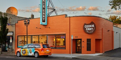 A delivery van has fleet graphics in orange and blue and is parked in front of a commercial building.

