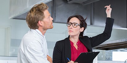In a commercial kitchen, a woman with a clipboard points up as a chef looks on.
