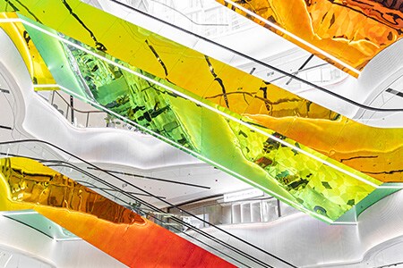 Shopping de vários níveis com paredes brancas e escadas rolantes suspensas cobertas com os refletivos e coloridos Acabamentos para Vidro Dicroico 3M em tons de vermelho, amarelo, laranja e verde.
