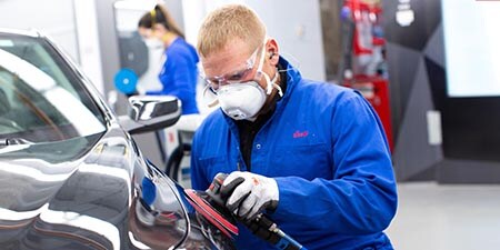 Um trabalhador utilizando uma lixadeira roto-orbital na carroceria de um automóvel em uma oficina.
