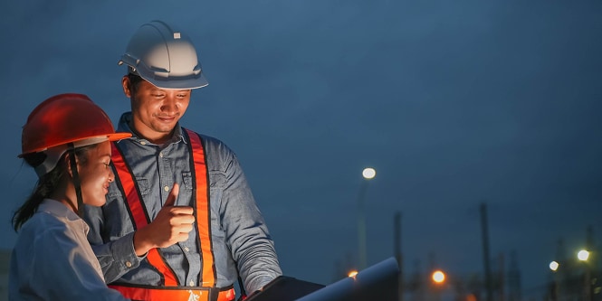 Pessoal da obra com capacetes e coletes refletivos revisa os planos ao anoitecer. 
