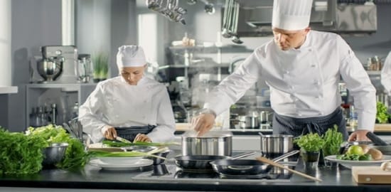 Três chefes preparam comida em uma cozinha comercial.
