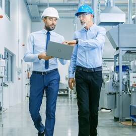 Dois funcionários de uma fábrica caminhando e discutindo o trabalho com um laptop na mão.