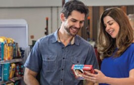 Duas pessoas em um ambiente de loja, observando juntas uma caixa de produto que seguram nas mãos. As faces estão desfocadas. Há prateleiras com itens ao fundo.
