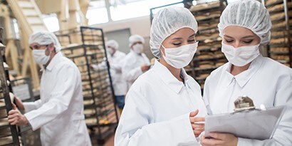 Trabalhadores usando máscaras e toucas de cabelo conferem em uma cozinha comercial.
