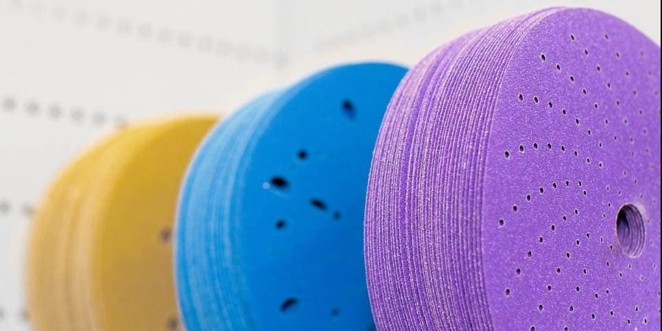 Variedade de discos e rolos abrasivos 3M coloridos (azul, roxo, amarelo) organizados sobre uma bancada vermelha em uma oficina.
