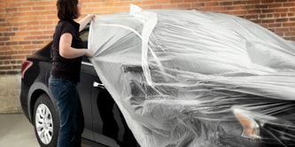 Pessoa cobrindo um carro preto com um filme plástico transparente de proteção em ambiente de oficina com parede de tijolos.
