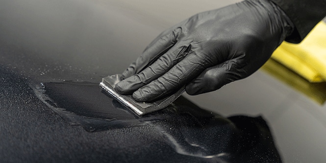 Uma pessoa usando luvas pretas está aplicando um revestimento em uma superfície usando um pequeno aplicador retangular, espalhando um líquido uniformemente por toda a área.
