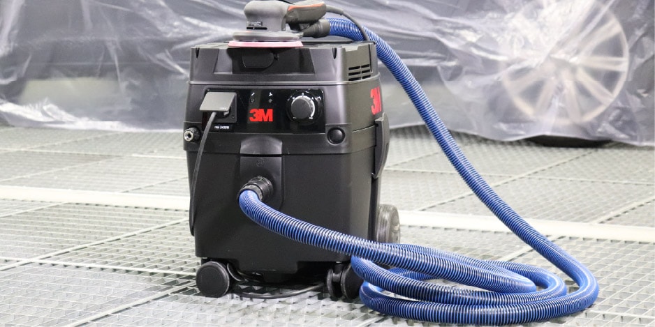 Extratora de pó preta da 3M com mangueira azul conectado a uma lixadeira, posicionado sobre piso de oficina com um carro ao fundo coberto por plástico de proteção.
