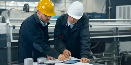 Duas pessoas usando capacetes olham para um tablet sobre uma mesa.
