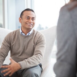 Man in tan shirt sitting in a chair conversing with a colleague. 