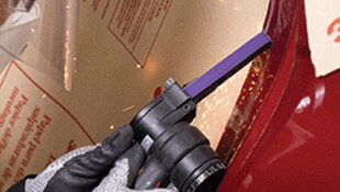 Uma mão com luva utilizando uma ferramenta elétrica portátil com uma correia abrasiva estreita sobre uma superfície metálica, produzindo pequenas faíscas
