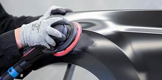 Trabalhador usando luvas de proteção utilizando uma lixadeira orbital com um disco abrasivo para nivelar um painel preto de veículo.
