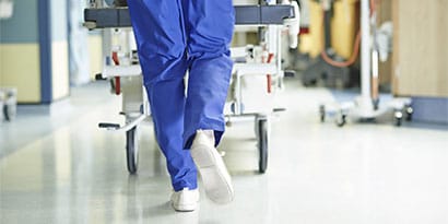 Health care provider’s feet walking away with gurney wheels in the background.

