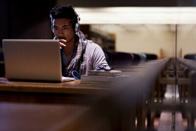 Imagem de um jovem usando fones de ouvido, sentado diante de um notebook, com livros ao lado, em um ambiente semelhante à uma biblioteca.
