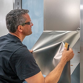 Homem instalando um acabamento prata metálico 3M DINOC na porta do elevador.