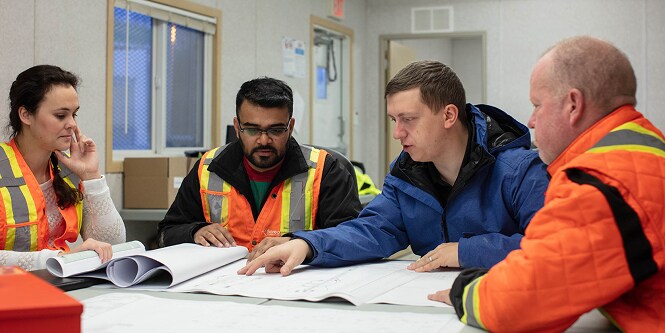 Equipe de construção analisa plantas sobre uma mesa dentro do escritório da obra.
