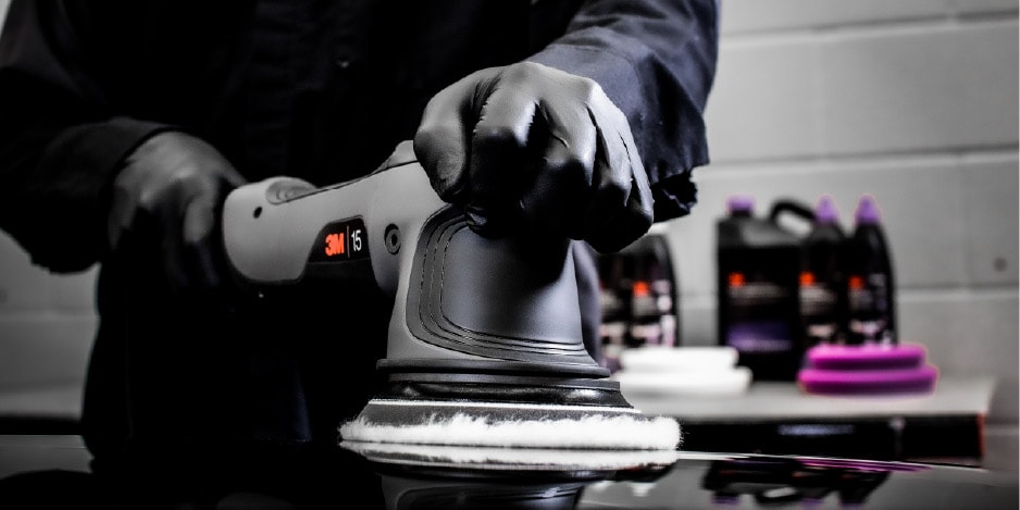 Pessoa utilizando uma politriz roto orbital 3M com luvas pretas para polir a superfície de um veículo, com produtos de polimento 3M ao fundo.
