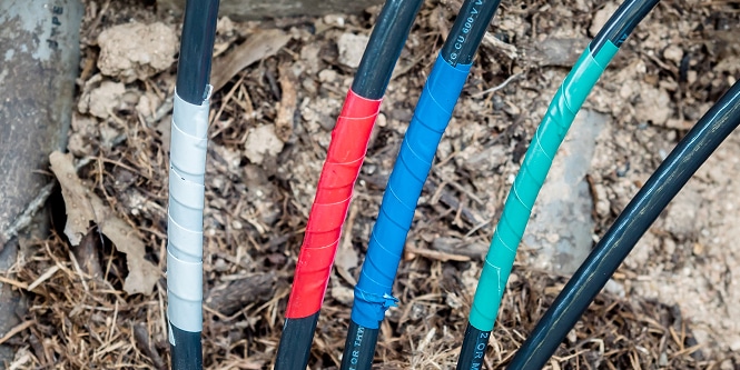 Quatro cabos externos envolvidos com fitas de cores diferentes - prata, vermelho, azul e verde - saindo do solo em uma área de terra e mulch.
